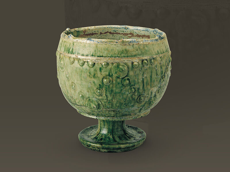 唐代．綠釉高足杯 此器呈歛口、深腹、高足之狀，表面壓印乳丁紋，施綠釉，是模仿外來金銀器的品類。（圖一）